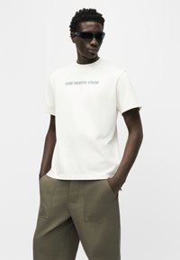 Άντρας που φοράει μαύρα γυαλιά ηλίου, λευκό μπλουζάκι The North Face και χακί παντελόνι, στέκεται με τα χέρια στις τσέπες.