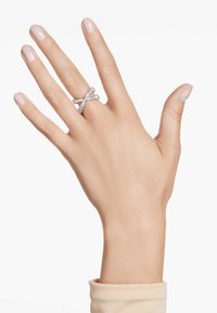 Anillo de plata con bandas entrelazadas, adornado con múltiples pequeñas piedras brillantes. Lleva en una mano con uñas pulidas de color claro.