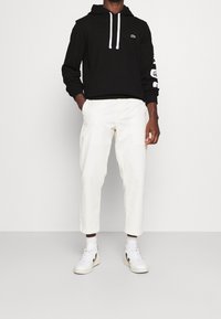 Felpa nera con cappuccio e cordini bianchi, con logo sul petto, abbinata a pantaloni bianchi a gamba affusolata e sneakers bianche con accenti neri.