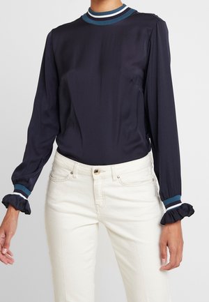 Frau trägt eine marineblaue Bluse mit langen Ärmeln, Rüschen an den Manschetten und einem gestreiften Kragen, kombiniert mit hoch taillierten weißen Jeans.