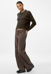 Mörkgrön kort tröja med krage och treknappslåsning, kombinerad med bruna jeans med vida ben och svarta skor. Mjuk tygtextur.