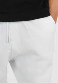 Bílé tepláky s bočními kapsami, stahovacími šňůrkami v pase a decentním logem. Tkanina vypadá měkce a pohodlně.