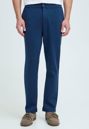 Mężczyzna noszący granatowe, casualowe spodnie z guzikiem i elastycznym pasem, w połączeniu z jasnobrązowymi zamszowymi butami.