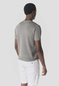 Man met kort krullend haar, gekleed in een grijs kort mouw breiselshirt en witte spijkershorts, staand met de rug naar de camera op een effen achtergrond.