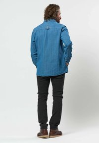 Homme aux cheveux ondulés vu de dos, portant une chemise en jean bleue, un pantalon noir et des chaussures marron sur un fond blanc uni.