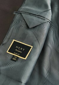 Giacca slim fit in satin grigio scuro con texture ondulata. Presenta un'etichetta in tessuto nera con rifiniture dorate che indica "NEXT Occasion", taglia 40R.