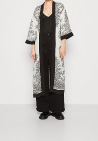 Długi, kimono-stylowy płaszcz z przezroczystej białej tkaniny w czarne wzory paisley, z czarnymi wykończeniami i rękawami o długości trzech czwartych.