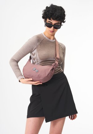 Helle rosa Schultertasche aus weichem Stoff, runde Form, mit zwei grauen Knöpfen und einem breiten Gurt. Vor einem neutralen Hintergrund getragen.