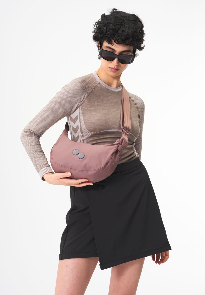 Helle rosa Schultertasche aus weichem Stoff, runde Form, mit zwei grauen Knöpfen und einem breiten Gurt. Vor einem neutralen Hintergrund getragen.