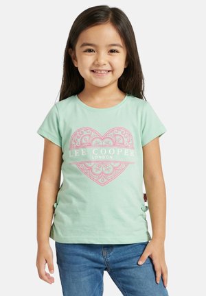 Jeune fille souriante, portant un t-shirt vert menthe avec un cœur décoratif rose et le texte « Lee Cooper London », associé à un jean bleu.
