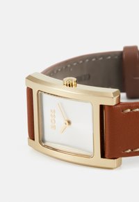 Montre rectangulaire dorée avec un cadran blanc, un bracelet en cuir brun et une couronne texturée. Le logo "BOSS" est affiché sur le cadran.