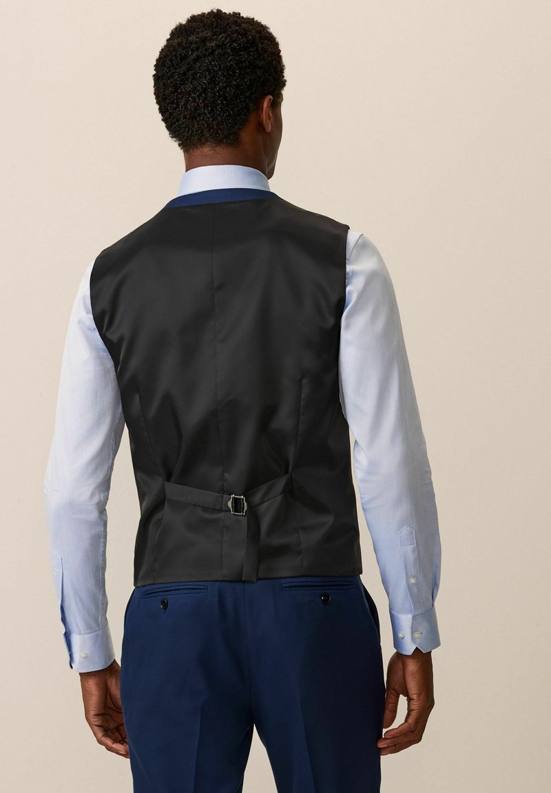 Chaleco negro con un acabado suave, con una hebilla en la parte posterior y llevado sobre una camisa azul clara con botones en contraste.