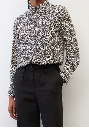 Person trägt ein schwarz-weißes, langärmeliges Hemd mit Blumenmuster, das in hoch taillierte schwarze Hosen gesteckt ist, die Hände in den Taschen.