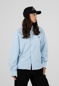 Jeune femme aux longs cheveux foncés porte une casquette noire, une chemise rayée bleu et blanc à boutons, et un pantalon noir, regardant vers sa droite.