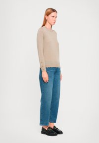 Beige Langarmpullover mit Rundhalsausschnitt und geknöpften Manschetten, kombiniert mit hochgeschnittenen blauen Jeans und schwarzen Slippern. Schlichtes, figurbetontes Design.