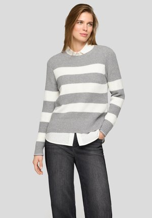 Woman wearing a gray and white striped sweater over a white collared shirt, paired with dark jeans, standing with one hand in pocket.
