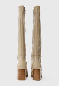 Knöchelhohe beige Stiefel aus Kunstleder mit glatter Textur, seitlichem Reißverschluss und gestapeltem Holzabsatz. Sie verfügen über ein minimalistisches Design und klare Linien.
