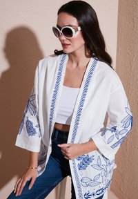 Valge kimono-tüüpi jakk siniste lillede tikandiga, 3/4 pikkuses varrukatega ja kontrastse sinise äärisega. Kandu valge särgi peal.