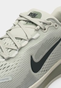 Chaussure de course beige clair avec un design complexe en mesh, logo Nike swoosh noir et lacets beiges noués sur la languette.