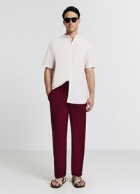 Camicia beige chiaro a maniche corte con colletto, abbinata a pantaloni larghi color bordeaux e sandali neri. Modello con occhiali da sole scuri.