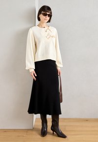 Pull crème avec des manches bouffantes et un détail en nœud, associé à une jupe noire côtelée et des bottes en cuir marron jusqu'au genou. Sac à main marron inclus.