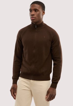 Mand iført en mørkebrun sweatjakke med lynlås, ribstrikket krave og manchetter, kombineret med lyse beige bukser, stående foran en ensfarvet baggrund.