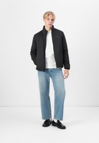 Svart polstret jakke med høy krage, brukt over en hvit skjorte, kombinert med lyseblå rette jeans og svarte sko.