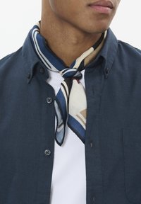 Camisa azul marino de botones, con una camiseta blanca debajo, acompañada de una bufanda de seda estampada en crema, azul y marrón, atada al cuello.