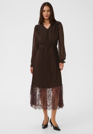 Femme portant une robe midi marron foncé à manches longues avec des plis, un ourlet en dentelle, des boutons sur le devant et une ceinture à la taille, assortie à des talons noirs.
