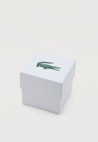 Μικρό λευκό κουτί με υφή, με πράσινο λογότυπο κροκοδείλου στο κέντρο του καπακιού, τοποθετημένο σε λευκή επιφάνεια.