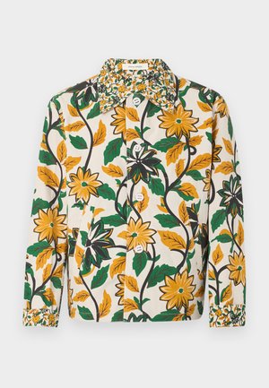 Homme portant un t-shirt blanc, une veste fleurie jaune et verte, un pantalon large marron foncé et des chaussures marron foncé, debout devant un fond uni.