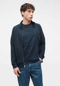 Νεαρός άνδρας που φοράει ένα μπλε σκούρο μπουφάν με φερμουάρ πάνω από ένα μπλε σκούρο μπλουζάκι και μπλε τζιν, στέκεται με το ένα χέρι στην τσέπη, κοιτώντας προς τα αριστερά του.