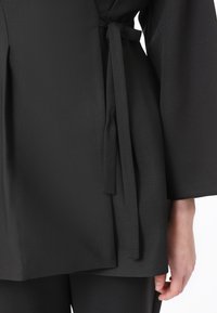 Top in tessuto nero con cintura annodata in vita, caratterizzato da maniche larghe e una texture liscia. Il tessuto cade in modo elegante.