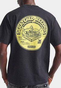 Camiseta de algodón negra con un gráfico de un disco de vinilo amarillo que incluye un gato y texto. El diseño incluye detalles como listas de canciones y un logotipo.