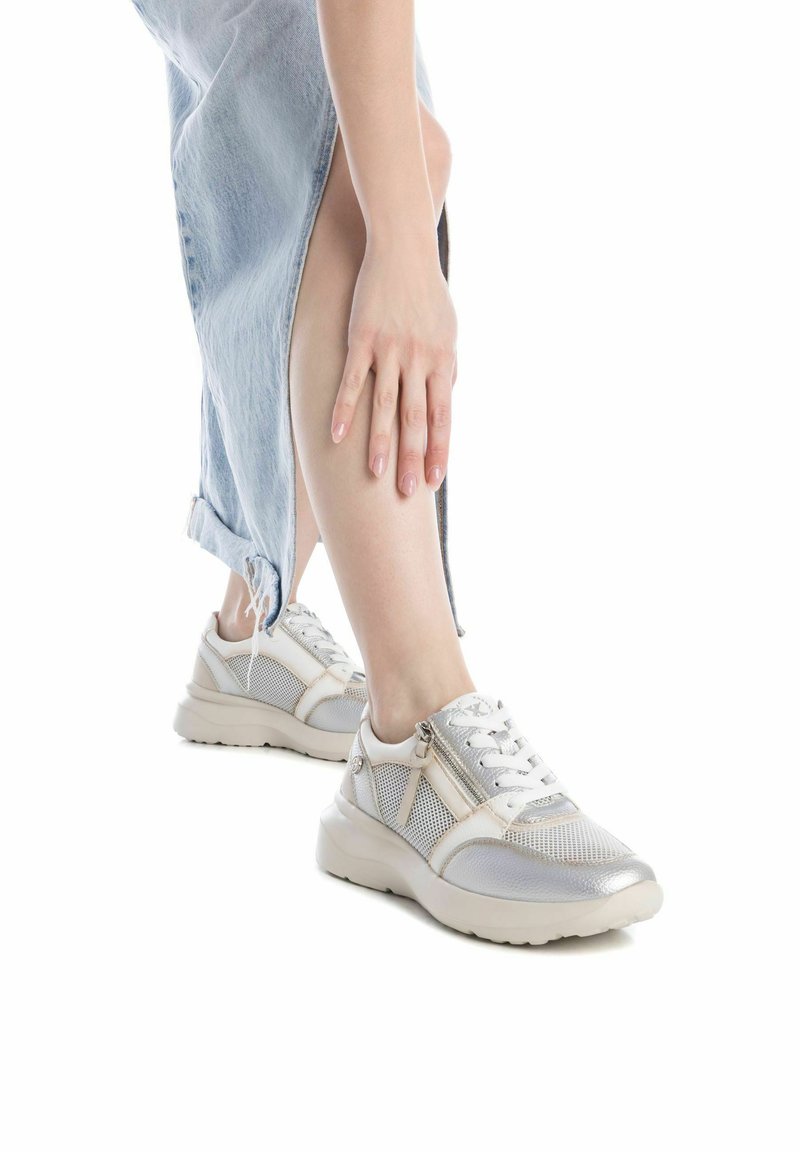 Zapatillas deportivas blancas y plateadas con detalles de malla, puntera redonda, suela gruesa y acentos de cremallera. Llevadas con una falda de mezclilla clara.