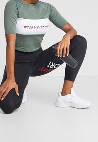 Camiseta atlética de manga corta verde con el logo blanco y rojo "TOMMY SPORT", combinada con leggins negros que tienen texto rojo y blanco, y zapatillas blancas.