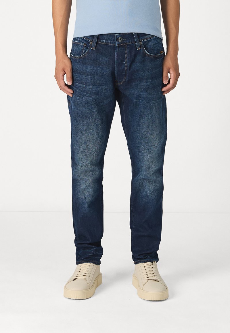 Donkerblauwe denim jeans, slim fit, vijf-pocket ontwerp, subtiele vervaging, gecombineerd met beige sneakers, gladde textuur.