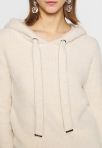 Beżowa bluza z kapturem o miękkiej fakturze, prążkowanym wzorze i sznurkami zakończonymi metalowymi zakończeniami, typowa dla stroju codziennego.