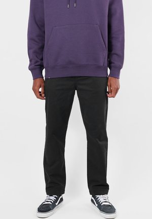 Personne portant un sweat à capuche violet, un pantalon noir et des baskets noir et blanc, debout devant un fond clair uni.