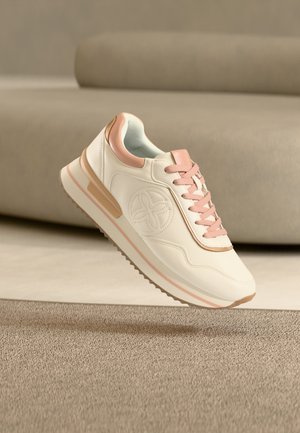 Λευκό και ροζ αθλητικό παπούτσι με σχεδιασμό κορδονιών και κυκλικό λογότυπο, τοποθετημένο στον αέρα πάνω από μπεζ χαλί και καναπέ στο φόντο.