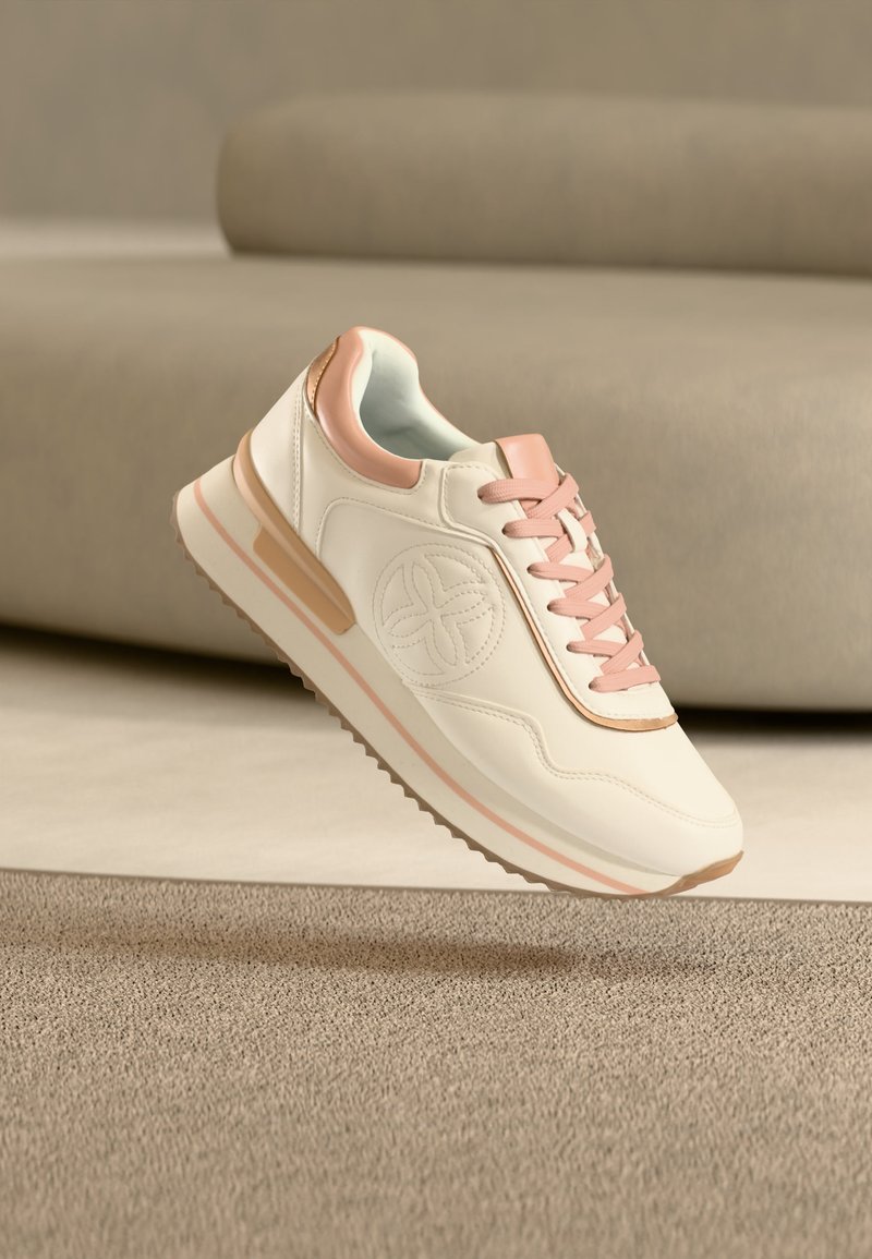 Λευκό και ροζ αθλητικό παπούτσι με σχεδιασμό κορδονιών και κυκλικό λογότυπο, τοποθετημένο στον αέρα πάνω από μπεζ χαλί και καναπέ στο φόντο.