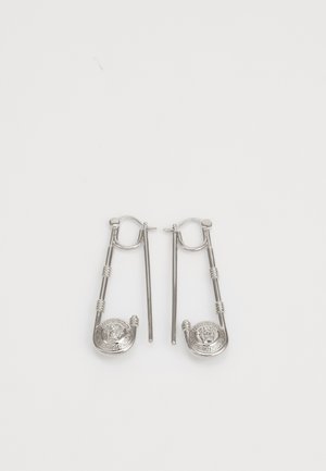 Brincos de prata em forma de clipes de segurança, com um medalhão redondo com um design de leão na parte inferior e padrões intrincados ao longo do comprimento.