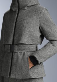 Šedá herringbone bunda s cropped strihom, kapucňou a čiernym zipsom. Obsahuje opasok v páse a bočné vrecká. Hladká textúra.