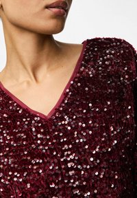 Burgunderfarvet paliettop med V-hals; har en tekstureret overflade og glat stofkant langs halsen.