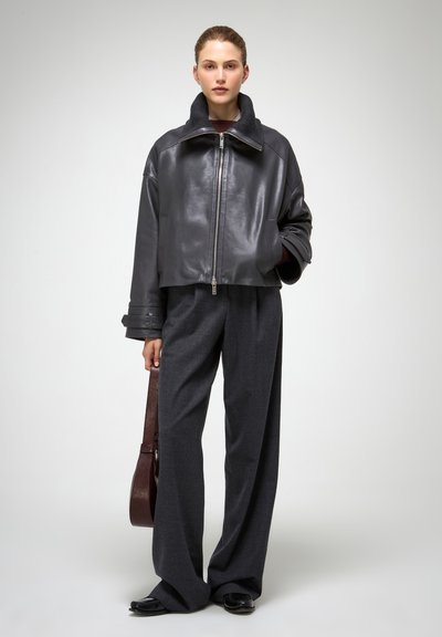 Giacca cropped in pelle nera con colletto ampio, abbinata a pantaloni grigi a gamba larga. La modella tiene una borsa marrone, in piedi su uno sfondo neutro.