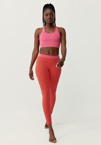 Rosa sportbh en koraalkleurige legging, gladde textuur, aansluitend ontwerp, met contrasterende tailleband. Model loopt blootsvoets op een neutrale achtergrond.