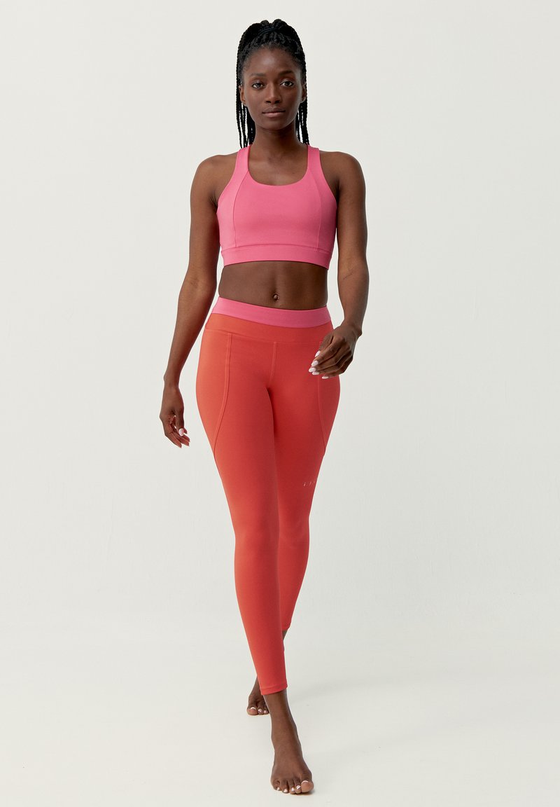 Rosa sportbh en koraalkleurige legging, gladde textuur, aansluitend ontwerp, met contrasterende tailleband. Model loopt blootsvoets op een neutrale achtergrond.