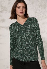 Langsleeve groene top met een geometrisch wit en teal patroon, voorzien van een V-hals ontwerp en een zachte textuur. Geschikt voor casual wear.