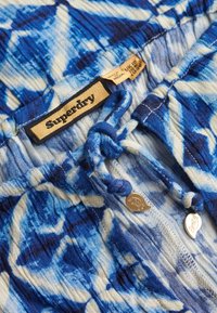 Tejido azul y blanco con patrón, con cordón de ajuste y etiqueta de Superdry; presenta material texturizado y acentos metálicos.