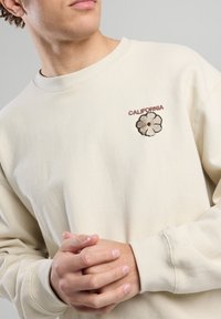 Sudadera de algodón beige con cuello redondo, que presenta el texto "CALIFORNIA" y un diseño de bordado floral en el pecho. Tejido texturizado.